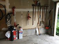 Wide view of wall-mounted garden tools including shovels, rakes, pitchfork, saw, axe, and hanging power tools (leaf blower and hedge trimmer).