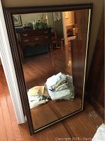 Framed rectangular wall mirror standing on the floor reflecting a wooden floor, furniture, and a pile of folded linens.