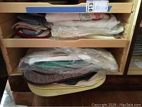 Shelf with stacked folded linens and tablecloths, some wrapped in plastic, in various colors including white, cream, and muted tones.