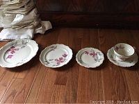 Full set showing various plate types and tea cups with saucers arranged on wooden floor with linens folded in background.
