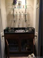 Wide view of bar area with shelves of glassware, glasses around sink, decanters, mixer blender on counter, silver-plate cups, and glass cabinet below with additional glasses and blender base.
