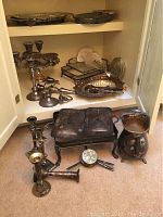 Wide shot of various silver-plated items including candlesticks, trays, fondue set, cake plate, and pitcher arranged on shelves and floor