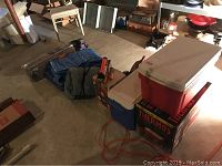 Wide shot of camping and outdoors items including coolers, sleeping bag, tarps, and tent bag in a basement setting.