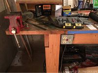 Left side of workbench showing the attached red metal vise mounted on the corner and the thick wooden surface with various tools on top.