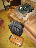 Multiple vintage luggage items arranged on chairs and carpet including green soft bag, large brown suitcase, small brown case, and wicker picnic basket with contents visible.