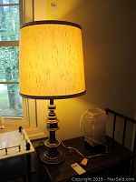 Brass colored table lamp with beige textured cylindrical shade lit and placed on wooden surface near window.