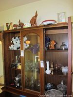 Wide view of wooden display cabinet with various ceramic and glass figurines including horses, owls, wooden elephants, and glassware inside