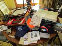Plastic model tank on wood board, hats with military and NRA logos, framed prints, books and pamphlets on round table with sunlight