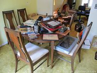 View of round wooden dining table with five wooden chairs with padded seats around it. Items have visible wear and scratches. Table is cluttered with miscellaneous items.