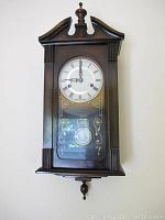 Front view of dark wood pendulum wall clock with glass door, white clock face with Roman numerals and decorative gold patterns on the glass.