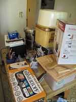 Overview of kitchen items on table including Hamilton Beach coffee maker in box, vintage tins, cutting boards, pens, and more