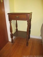 Side table showing turned legs, drawer with metal handle, and lower shelf, placed against a wall.