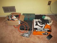 Overview of lot showing Black and Decker circular saw box, metal toolbox, gloves, hand tools, and power tools with electrical cords