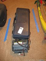 Closed black plastic carrying case with hydraulic jack inside, battery charger placed in front with attached power and battery clips cords.