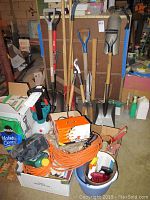 Wide view of garden tools including shovels, post hole digger, electrical cords, sprayer, bags of potting mix, and garden accessories in buckets.