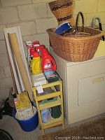 Overall view of metal shelving holding household chemicals such as laundry detergent, baskets on top, and other items including a blue bucket below.