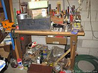 Front view of the workbench with one drawer and visible orange vise on the side, surrounded by miscellaneous tools and supplies not included in the lot.