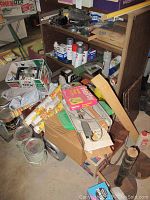 Image of a workspace floor and low shelves cluttered with various items such as wallpaper rolls in original packaging, small boxes, spray cans, automotive products, and hardware supplies.