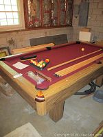 Side angle photo of the wooden framed pool table with burgundy felt, showing pool cues, balls in a triangle, brush and chalk on the table surface.
