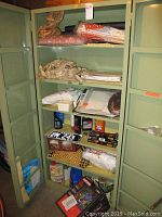 Metal cabinet containing various items including a Kansas license plate (EM 243), rolls of material, and packaged macrame supplies.