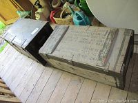 Photo showing two wooden trunks with address lettering on the top and side surfaces, placed on wooden floorboards.