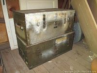 Front and top view of two stacked trunks showing metal surface with rivets, latches, and visible wear including rust, scratches, and faded markings.