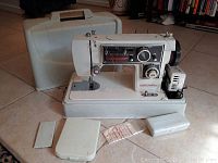 Front and side view of white Dressmaker sewing machine showing stitch selector panel with multiple stitch types and dial controls. Machine mounted on base with foot pedal, plastic case, and two small plastic accessory boxes visible.