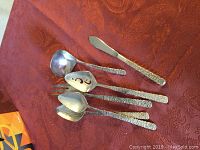 Photo of 5 silver plated serving pieces on reddish textured cloth, showing the textured handles and pierced decoration on one spoon.