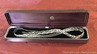 Sterling silver braided necklace inside an open Cortesis branded jewelry box with maroon velvet lining, displayed on a red patterned fabric surface.
