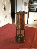 Photo showing a copper and brass miners lamp on a red tablecloth in a kitchen setting. The lamp has a cylindrical shape with a glass enclosure and a decorative emblem on the body.