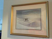 Framed print showing two figures carrying skis walking over snowy dunes under a cloudy sky. The image includes signature and identifying marks.
