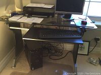 Black glass computer desk with printer, monitor, keyboard, mouse, and computer tower underneath, placed near a window.