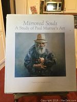Photo of the cover of a hardcover art book 'Mirrored Souls: A Study of Paul Murray's Art' showing a painting of an elderly man in blue holding a small dog.