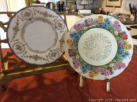Photo showing two vintage decorative ceramic teaplates on stands, one with beige scalloped edging and floral garlands, the other with colorful floral rim and scalloped edges.