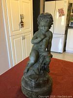 Full view of the 16-inch Cupid statue showing the figure sitting on a textured base, placed on a table in a kitchen setting.