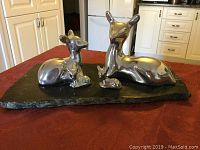 Polished aluminum sculptures of a deer family, an adult deer and a fawn, mounted on a natural edge black marble slab displayed on a red tablecloth in a kitchen setting.