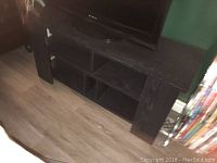 Black wooden TV stand on wood floor with four open storage shelves, some minor scratches visible on the top surface.
