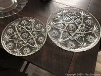 Two glass serving trays with detailed pinwheel patterns and scalloped edges, one larger than the other, shown on a wooden surface.