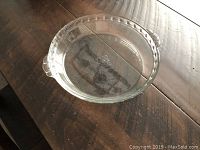 Top-down photo of clear glass pie dish with scalloped edges and handles on wooden surface.