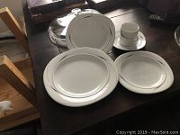 Photo showing the Mikasa Caviar place setting components arranged on a wooden table, including coffee cup, saucer, dinner plate, salad plate, and soup bowl with silver trim.