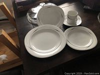 Photo showing Mikasa Caviar place setting items arranged on a dark surface: includes coffee cup and saucer, dinner plate, salad plate, soup bowl all white porcelain with silver or platinum trim