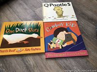 Photo showing three hardcover children's books arranged on a wooden floor: One Duck Stuck, QPootle5, Cowboy Kid.