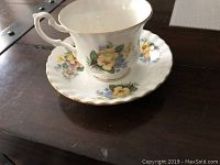 Bone china tea cup and saucer with yellow and purple flowers and gold trim edges shown from side view on table.