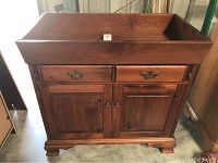 Front view of the solid wood dry sink showing two drawers with metal handles above two cabinet doors with wooden knobs. The wood has a light brown finish and is raised on bracket feet.