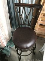 Photo of one barstool showing dark brown cushioned seat, ornate metal backrest, and metal frame with circular footrest