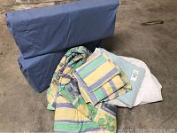 Photo showing twin reversible bed linen set components arranged on the floor including blue bolster-style pillows, striped comforter and sheet set, mattress pad, bed skirt, and white blanket.