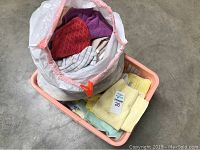Photo of a light pink plastic basket with assorted folded rags and a large white plastic bag filled with various cloth rags on a concrete floor.