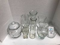 Seven clear glass vases and one ribbed glass jar with lid arranged in a white corner background.