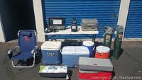 Photo showing various coolers, camping chairs, portable grills, and tent bags arranged outdoors against a blue storage unit door.