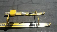Yellow water ski seat with attached dual skis, showing the side profile on pavement.